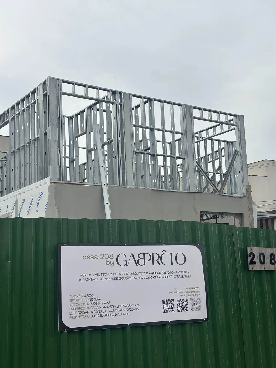 Tapume da obra com a placa da Casa 208 e a estrutura em Steel Framing ao fundo